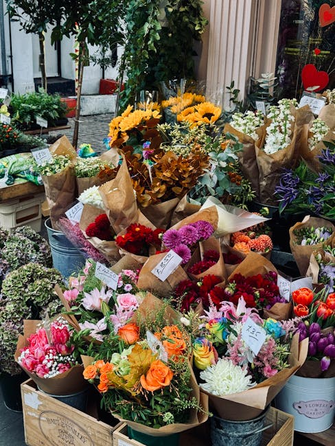 A vibrant display of fresh flower bouquets arranged on a wooden table outside a florist shop, featuring a variety of blooms including pink lilies, orange roses, purple irises, white chrysanthemums, re