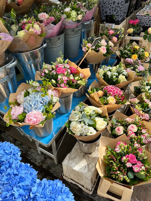 An outdoor display of fresh floral bouquets arranged in metal buckets and wrapped in brown paper, featuring a variety of flowers including pink roses, white carnations, pale yellow blossoms, blue hydr