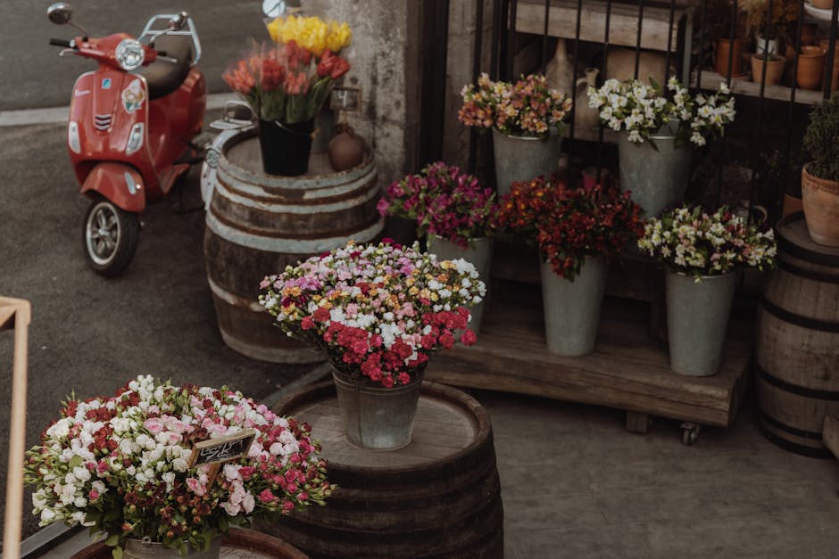 A display of fresh floral arrangements at a flower shop, featuring a variety of chrysanthemums and mixed flowering plants in metal buckets and on wooden barrels. The flowers include pink, white, yello