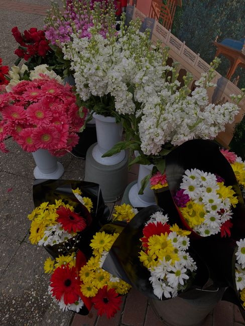 A colorful display of fresh flower bouquets and floral arrangements outside a shop, featuring pink chrysanthemums, white stock flowers, yellow daisies, and red gerbera daisies, all neatly wrapped in b