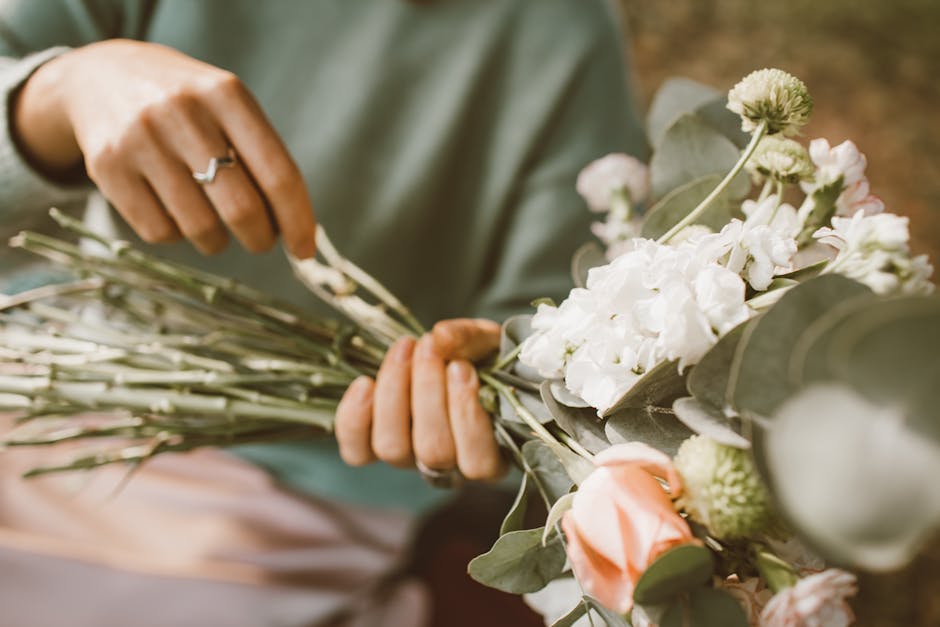 A person is holding a bouquet of fresh flowers, including white hydrangeas, soft pink roses, and green accents, wrapped in greyish-green foliage. The arrangement is being carefully assembled, with the