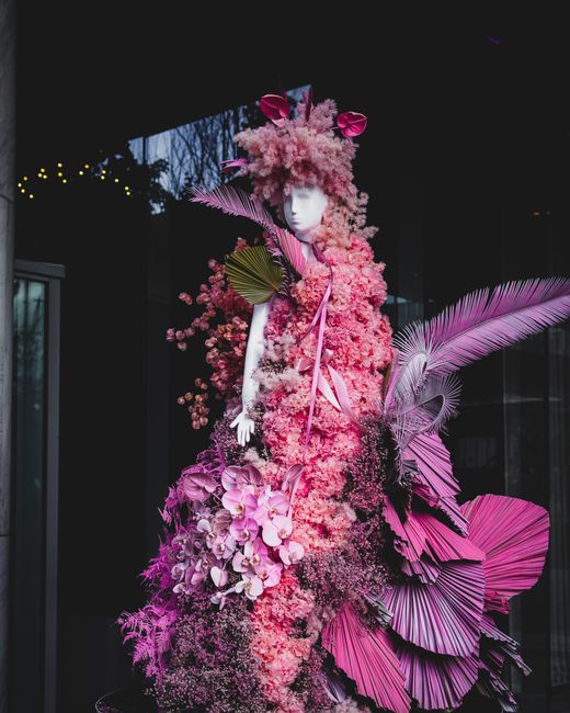A striking floral display featuring a mannequin adorned with an elaborate arrangement of pink flowers, including fluffy pink blossoms resembling cherry blossoms or hydrangeas, and cascading orchids wi