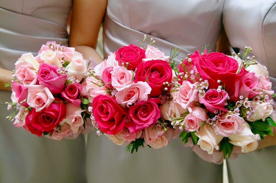 A floral arrangement featuring three coordinating bouquets of fresh roses in shades of pink, blush, and deep red, held by individuals dressed in neutral-colored clothing. The bouquets are composed of