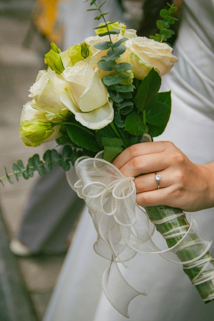 A bridal hand holding a fresh floral bouquet featuring creamy white roses, light green buds, and greenery including eucalyptus and eucalyptus-like leaves, wrapped with a delicate, sheer white ribbon.